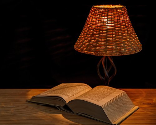 Person reading a book in a cozy room illuminated by a warm desk lamp